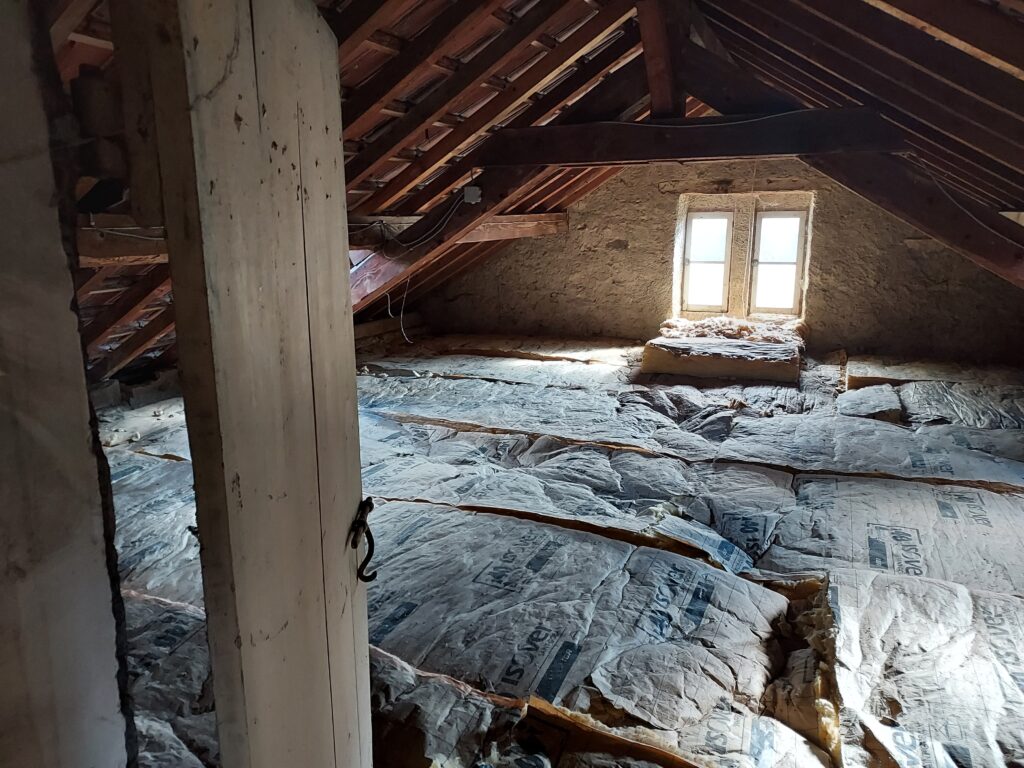 Grenier en cours d'isolation, avec de la laine de verre encore apparente qui est en train d'être posé au sol sous une charpente en bois apparente et une lumière naturelle entrant par une petite fenêtre à double battant.  - Agrandir l'image, fenêtre modale