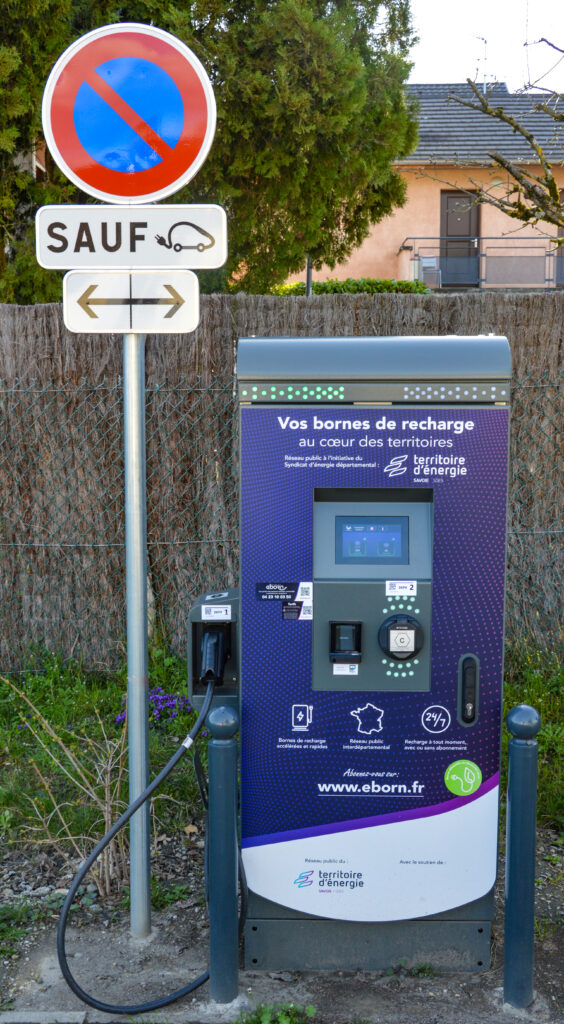 Photo d'une borne de recharge pour véhicules électrique, à côté d'un panneau de signalisation "stationnement interdit sauf véhicules électriques". 
En arrière-plan, une palissade et une maison masquée en partie par un arbre.  - Agrandir l'image, fenêtre modale