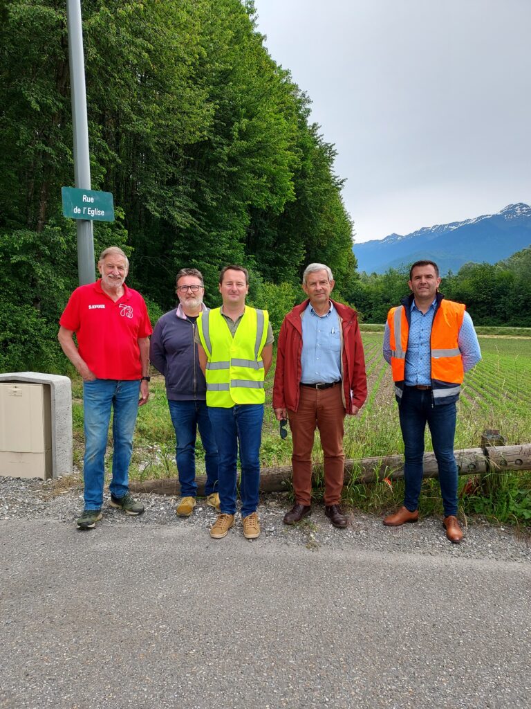 Cinq hommes, dont deux avec des gilets réfléchissant, sont debout en ligne au bord d'une route, devant un paysage verdoyant avec des arbres, une montagne au loin et un champ.  - Agrandir l'image, fenêtre modale