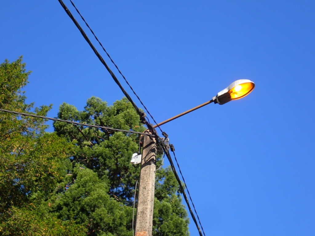Poteau électrique en béton avec plusieurs câbles attachés au sommet, surmonté d’un lampadaire allumé diffusant une lumière jaune. En arrière-plan, des arbres verts et un ciel bleu clair sans nuages. - Agrandir l'image, fenêtre modale