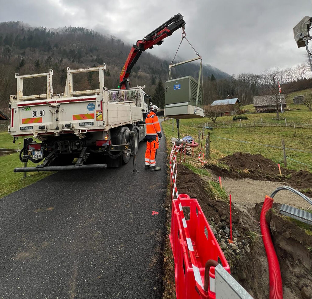 homme en combinaison réflechissante orange, devant un camion avec un bras mécanique qui soulève un post électrique au dessus d'une tranchée balisée par des barrières de chantier rouges et une rubalise rouge et blanche rayé. 
En arrière plan on voit le toit de deux maison, une montagne et un ciel gris.  - Agrandir l'image, fenêtre modale