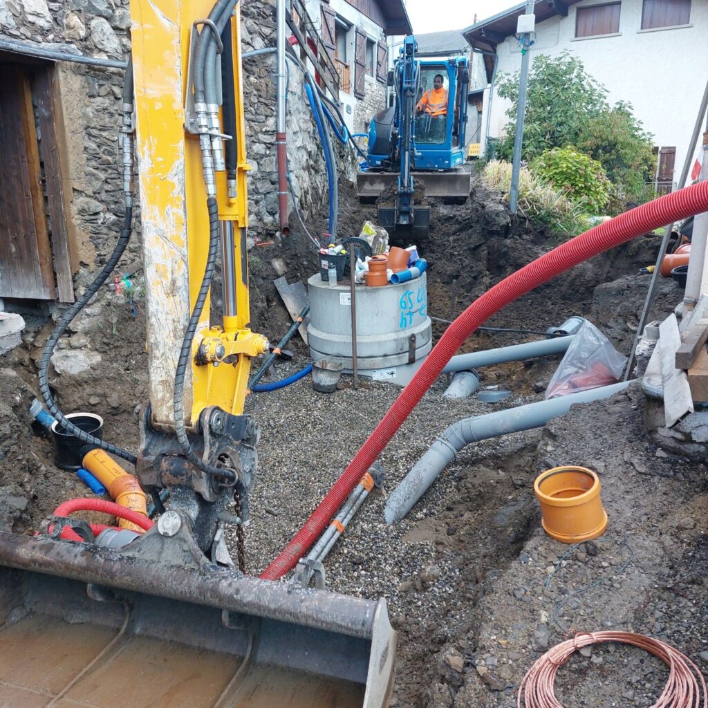 photo d'une tranché creusé à côté d'une maison en pierre seches. Un homme en tenue refléchissante orange est au commande d'une pelle mécanique bleu pour creuser une tranchée, dans laquelle se trouve des tuyaux gris et rouges. Au premier plan un bras de pelle mécanique jaune est posé. A l'arrière plan on voit deux maison au facade blanche et au volet en bois marron.  - Agrandir l'image, fenêtre modale